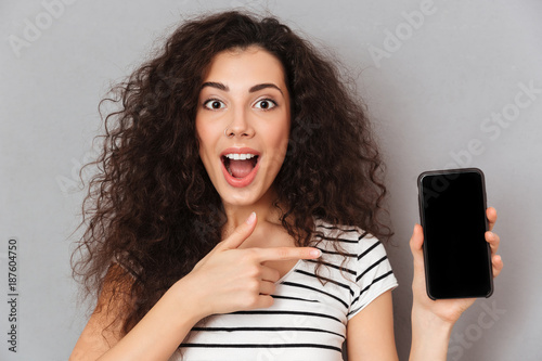 Close up photo of optimistic woman with ring in nose pointing index finger like advertising her smartphone meaning good choice against grey wall