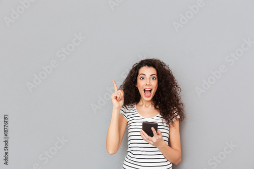 Smart female with mobile phone in hand keeping index finger pointed upwards expressing she has an idea or saying eureka over grey background