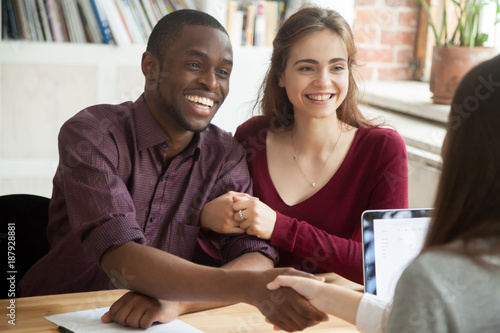 Smiling multiracial couple customers shaking hands with mortgage broker or financial advisor, happy attractive black and white family handshaking lawyer bank worker taking loan, making purchase deal