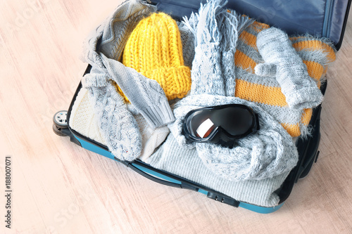 Open suitcase with warm clothes and ski goggles on wooden floor. Winter vacation concept