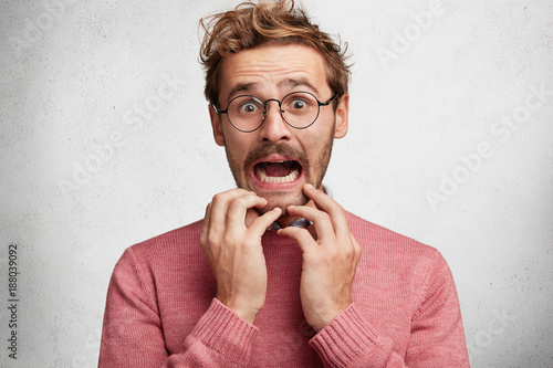 Worried scared fashionable man looks with terrified expression, keeps arms on chin, dressed in pink sweater, isolated over white background. Emotional bearded young guy receives unexpeted news