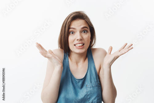 Portrait of funny good-looking european student girl with dark long hair in blue casual clothes looking in camera with confused expression, gesticulating with hands showing she can't answer question