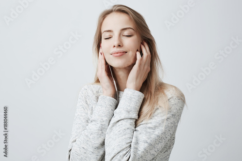 Close-up portrait of beautiful attractive European young woman in gray loose sweater, relaxing with closed eyes, listening to her favourite songs via white earphones, using music app.