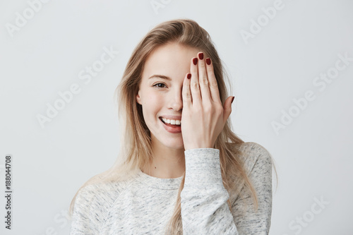 Happy smiling female with attractive appearance and blonde hair wearing loose sweater showing her broad smile having good mood closing her eye with hand, enoying to pose at camera. Happiness and joy