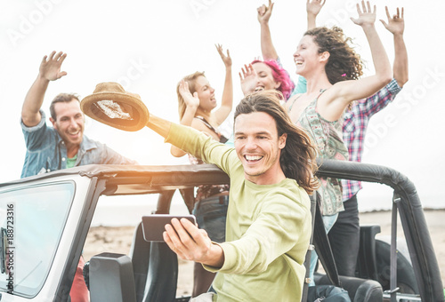 Trendy happy friends taking selfie with smartphone in desert on convertible jeep car - Travel people having fun together in excursion - Friendship and vacation concept - Focus on man's face with phone