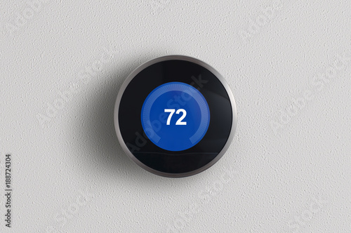 A simplistic photo of a round, modern, programmable digital thermostat in cooling mode, on a clean white wall. 