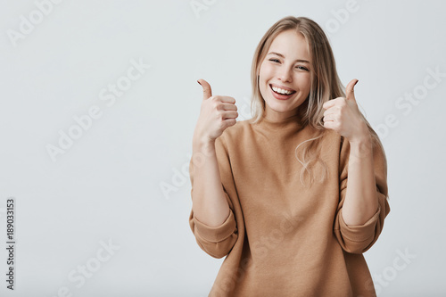 Portrait of fair-haired beautiful female student or customer with broad smile, looking at the camera with happy expression, showing thumbs-up with both hands, achieving study goals. Body language