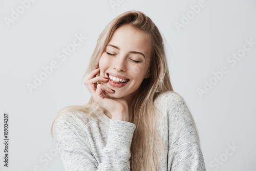 Beautiful female closing eyes because of joy, with long blonde hair and gentle smile. Cheerful woman holds hand under chin, has pleased expression of face. Face expression and positive emotions
