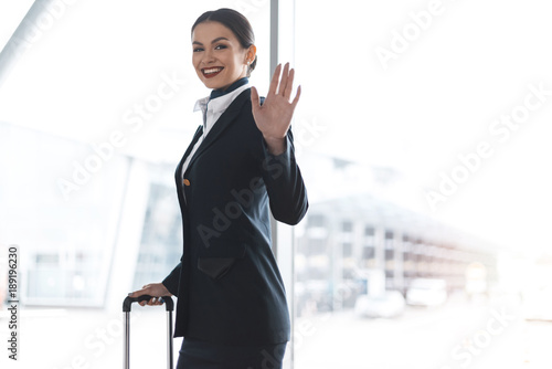 attractive young stewardess with suitcase waving at camera in airport