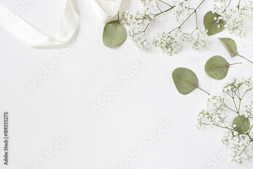 Styled stock photo. Feminine wedding desktop mockup with baby's breath Gypsophila flowers, dry green eucalyptus leaves, satin ribbon and white background. Empty space. Top view. Picture for blog.