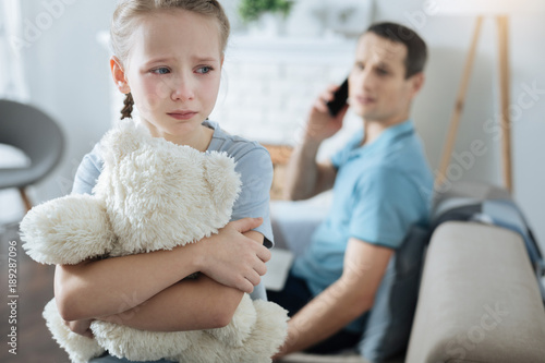 Poor child. Unhappy blue-eyed fair-haired little girl holding her toy and crying while her father talking on the phone