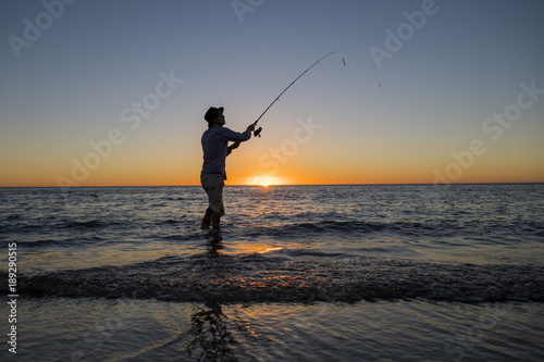 silhouette of fisherman with hat on the beach with fish rod standing on sea water fishing at sunset with beautiful orange sky in vacations