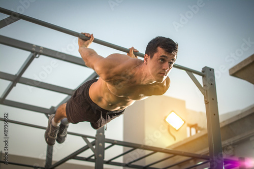 Young shirtless man doing exercises on horizontal bar outdoors. Calisthenics workout.