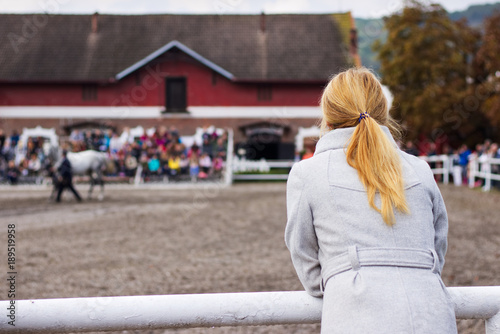 Blonde woman is watching a horse auction, English thoroughbred on a show. 