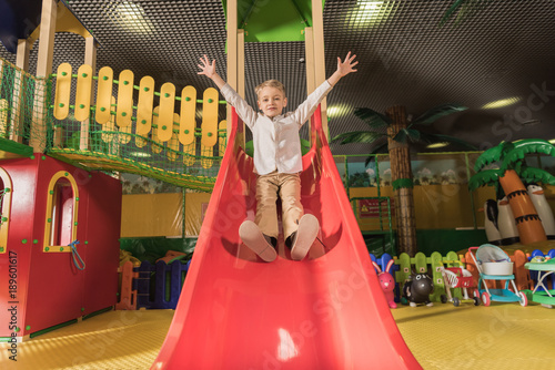 cute happy little boy with raised hands playing on slide in entertainment center