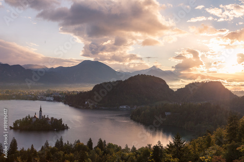 A neutral & pastel colored sky at Lake Bled. The sun is rising at Lake Bled uncovering the town in the distance. A thin layer of fog can be seen in the distance.