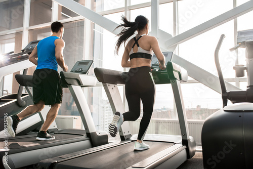 Full length portrait of two fit young people, man and woman, running on treadmills facing windows in modern gym, copy space