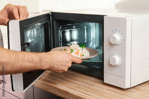 Man putting plate of rice with vegetables in microwave oven, closeup