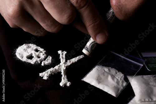 Drug dependence Kills. Junkie man sniffing a line of cocaine in the shape of a skull through a dollar, sachets with a dose. Black background. Narcotics concept.