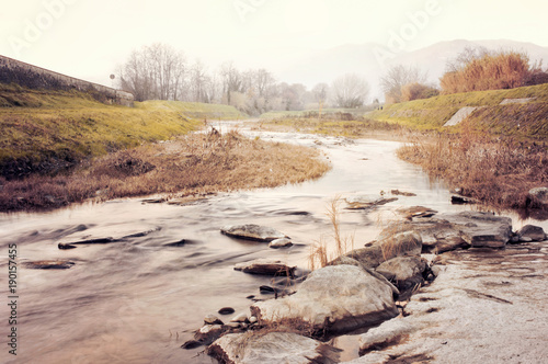 Panoramic view of the Pescia river, in the Italian countryside, with water and stones in the foreground and vegetation in the background, filtered with warm tones