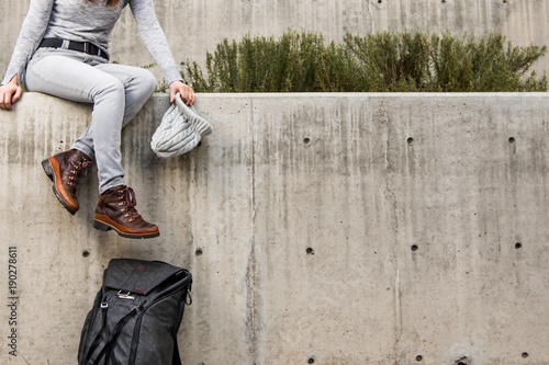 Woman dangling legs off the edge of a concrete ledge with grey beanie hat in hand and backpack leaning against wall. Lots of negative space to the right.