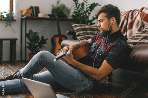 Young guitarist hipster at home sitting on the floor holding guitar listening earphones sound