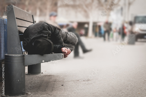 Poor homeless man or refugee sleeping on the wooden bench on the urban street in the city, social documentary concept, selective focus