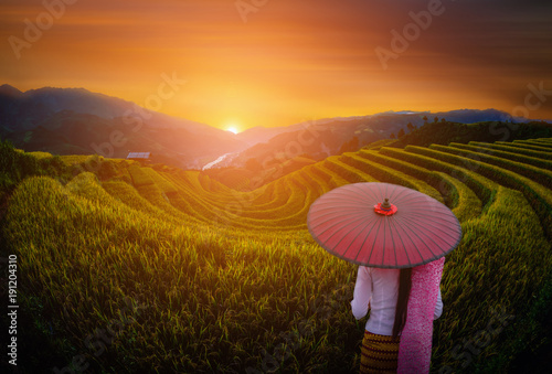 Woman holding traditional red umbrella on rice fields terraced with wooden pavilion at sunset in Mu Cang Chai, YenBai, Vietnam.