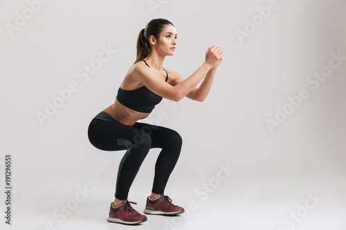 Image of sporty athletic woman in sneakers and tracksuit squatting doing sit-ups in gym, isolated over gray background