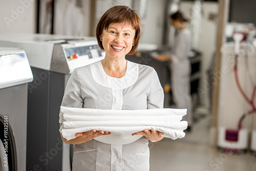 Portrait of a senior washwoman in uniform standing with bedclothes in the hotel laundry