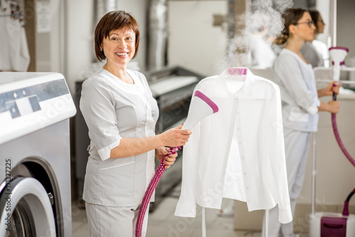 Portrait of a senior washwoman in uniform ironing up a white shirt with vapor machine in the professional laundry