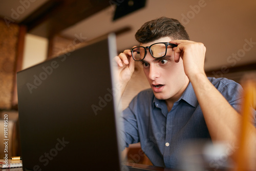 Shocked freelancer hipster man looks to laptop screen and can not believe unpleasant news. Pop-eyed frightened businessman trader raises one's glasses above his eyes. Trader monitoring stock exchange