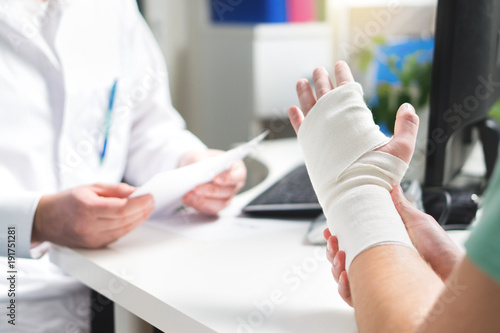 Injured patient showing doctor broken wrist and arm with bandage in hospital office or emergency room. Sprain, stress fracture or repetitive strain injury in hand. Nurse helping customer. First aid.