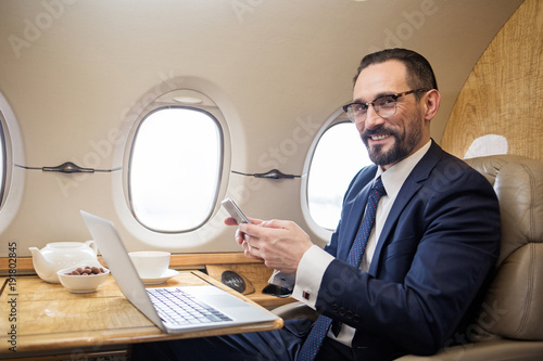 Portrait of contented diplomat sitting at tray table in airplane and holding cellphone in hands, he is looking at camera and smiling