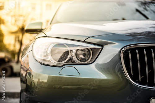 radiator grille and headlight of a blue car against the background of a blurred building with morning sun rays