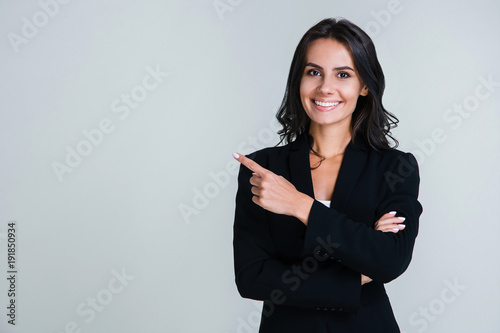 You need to look here! Beautiful young businesswoman pointing away and looking at camera with smile while standing against white background