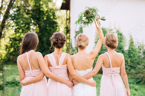 bride standing with her back and raised hand with a bouquet in the embraces of three bridesmaids in pink dresses transformer 