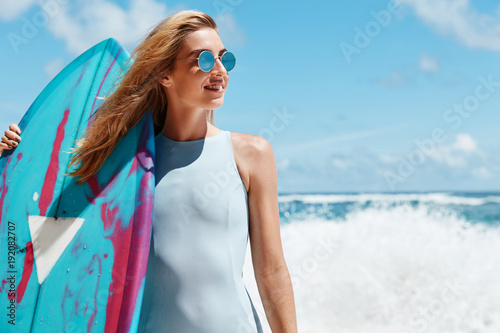 Slim fit female model poses in swimwear, holds kite board, stands against stromy ocean, has appropriate weather conditions for surfing, daydreams of driving, has pleased satisfied expression
