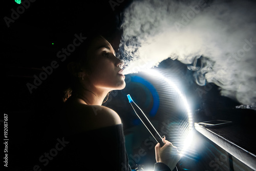 Young, beautiful girl smokes a hookah in the night club. Backlight LED. It produces smoke from his mouth. The pleasure of smoking.