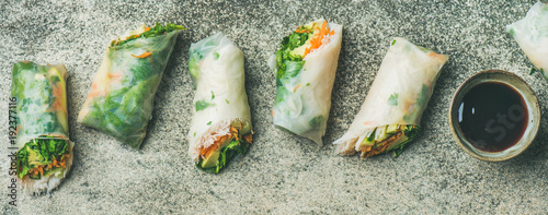 Helathy Asian cuisine. Flat-lay of vegan spring rice paper rolls with vegetables, soy sauce, chopsticks over concrete background, top view, wide composition. Clean eating, dieting, vegetarian food