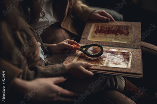 Child girl with woman in image of Sherlock Holmes sits and looks photoalbum with magnifier.