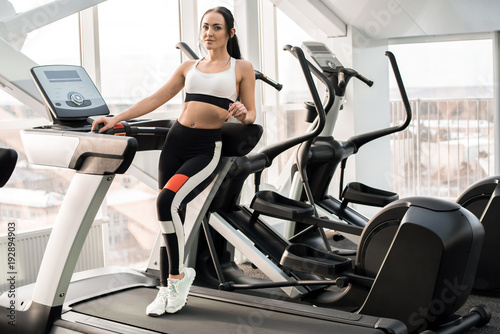 Full length portrait of fit young woman posing standing on treadmill in modern gym, copy space