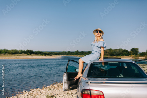 Young traveling woman in a blue dress is sitting by the car by the river
