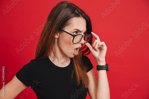 Photo of young outrageous woman wearing black t-shirt taking off glasses and looking aside with indignation or shock isolated over red background