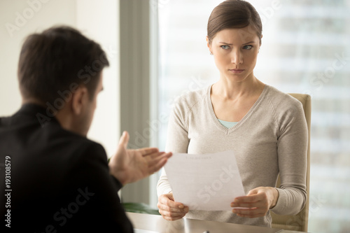 Millennial businesswoman with skeptical facial expression holding contract document and listening unconvincing offer of business partner. Suspicious terms of agreement, doubtful investor, fraud, scam