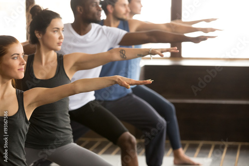 Group of young sporty afro american and caucasian people practicing yoga lesson, standing in Warrior two exercise, Virabhadrasana II pose, working out, indoor close up, studio