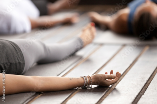 Group of young sporty people practicing yoga, lying in Corpse pose, Savasana exercise, working out, resting after practice, female hand with wrist bracelets close up, studio. Healthy lifestyle concept