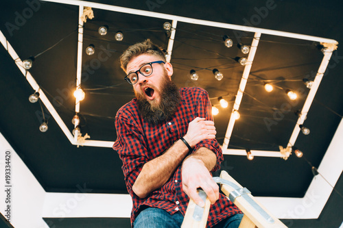 Hipster creative beard guy in eyeglasses balancing on construction worker climbing a ladder while working on home interior over black ceiling background. Anxiety unhappy expression concept.