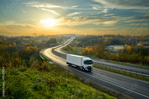 White truck driving on the highway winding through forested landscape in autumn colors at sunset