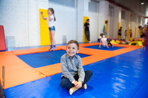 happy smiling little boy sitting on trampoline among jumping kids indoors